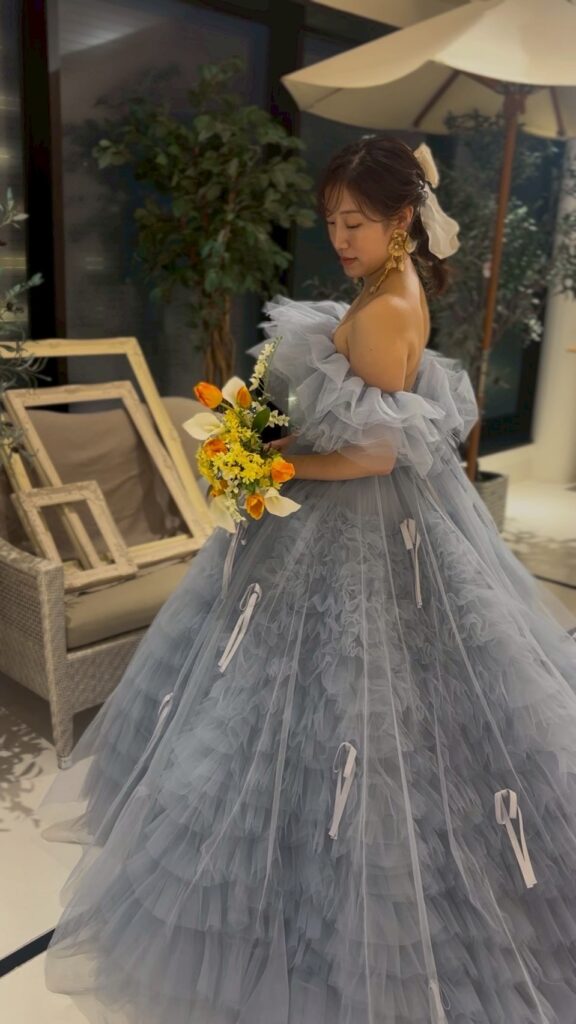 ガーデン風のスタジオにて、淡いブルーのフリルのドレスを着用し、ブーケを手にして振り返る新婦の全身ショット。上品でロマンチックに。