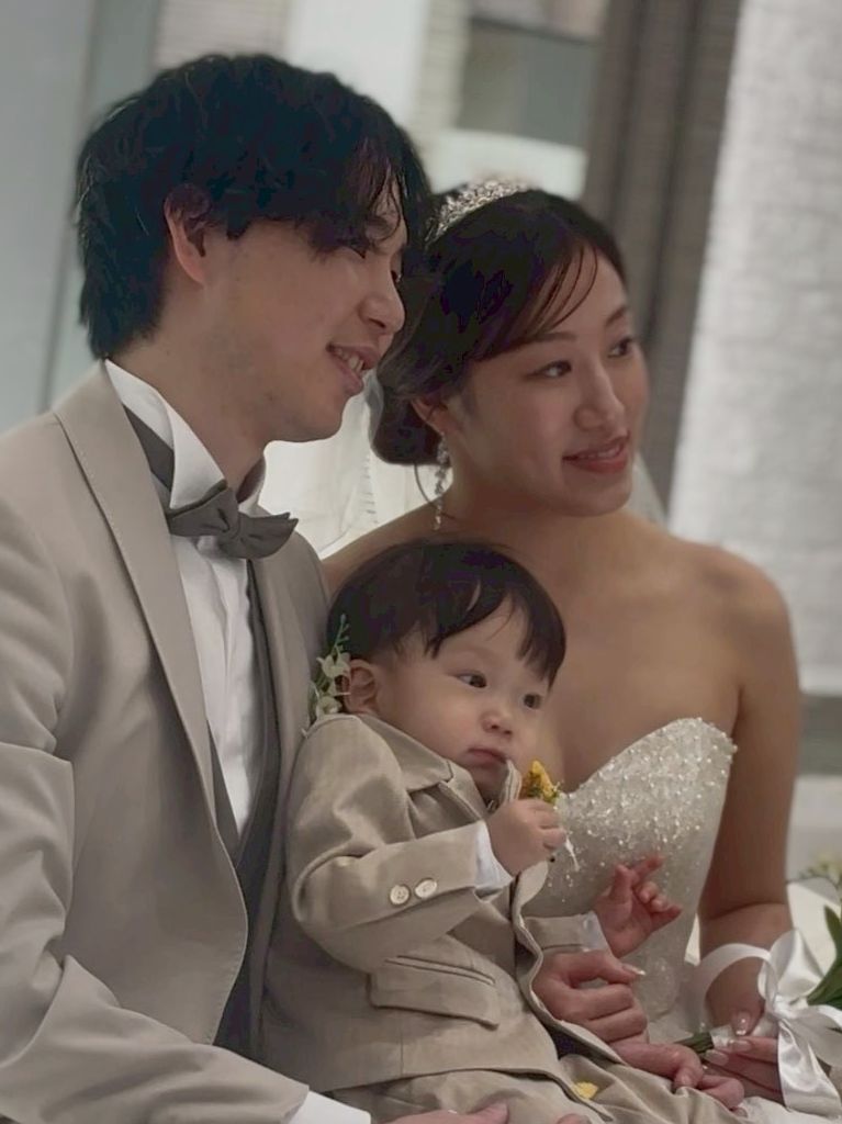 タキシード姿の新郎とウエディング姿の新婦が中央にいる赤ちゃんを優しく見つめ、3人で幸せそうに微笑むファミリーショット。