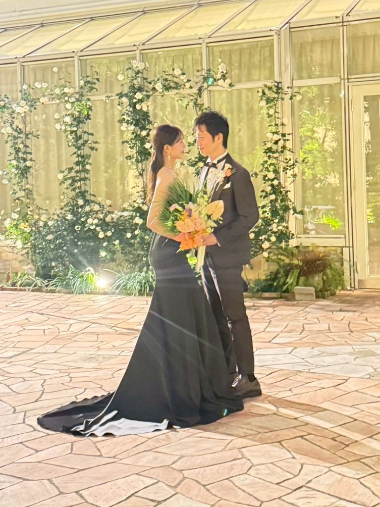 グリーンが美しい開放的な空間で、寄り添いあうタキシード姿の新郎と黒いドレス姿の新婦。